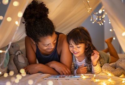 African mother and cute smiling girl using digital tablet while lying in illuminted tent in kid bedroom. Cheerful ethnic woman and lovely daughter on video call under a cozy hut. Lovely little girl with mom watching cartoon on digital tablet in bedroom.